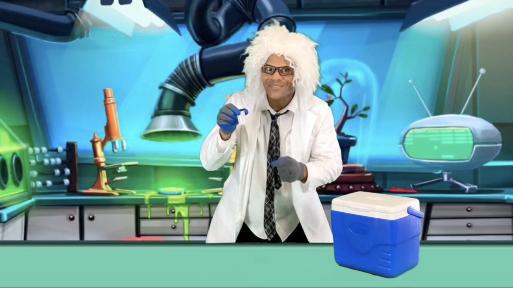 A Science Teller wears a white lab coat and white wig while standing in front of a cartoon laboratory backdrop. He holds a vial, ready to start an experiment.