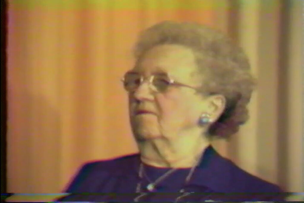 Video still capture of Irene Draper. Irene is an older woman with a gray perm, wearing glasses and opal earrings.