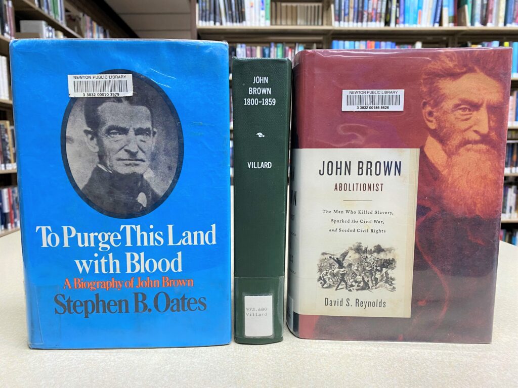 Three library books about John Brown are displayed on a table. Titles shown are "To Purge This Land with Blood," by Stephen B. Oates, "John Brown 1800-1859," by Oswald Villard, and "John Brown, Abolitionist," by David S. Reynolds.