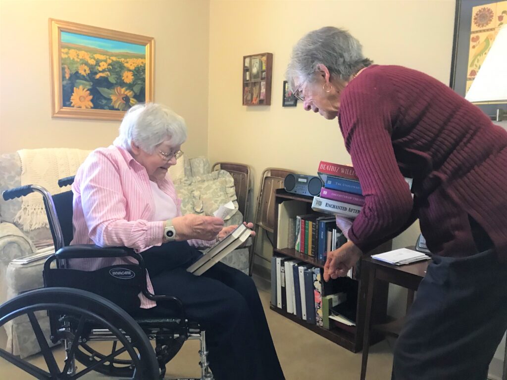 An ENLITE volunteer delivers books to an elderly library patron seated in a wheelchair inside her apartment.