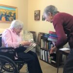 An ENLITE volunteer delivers books to an elderly library patron seated in a wheelchair inside her apartment.
