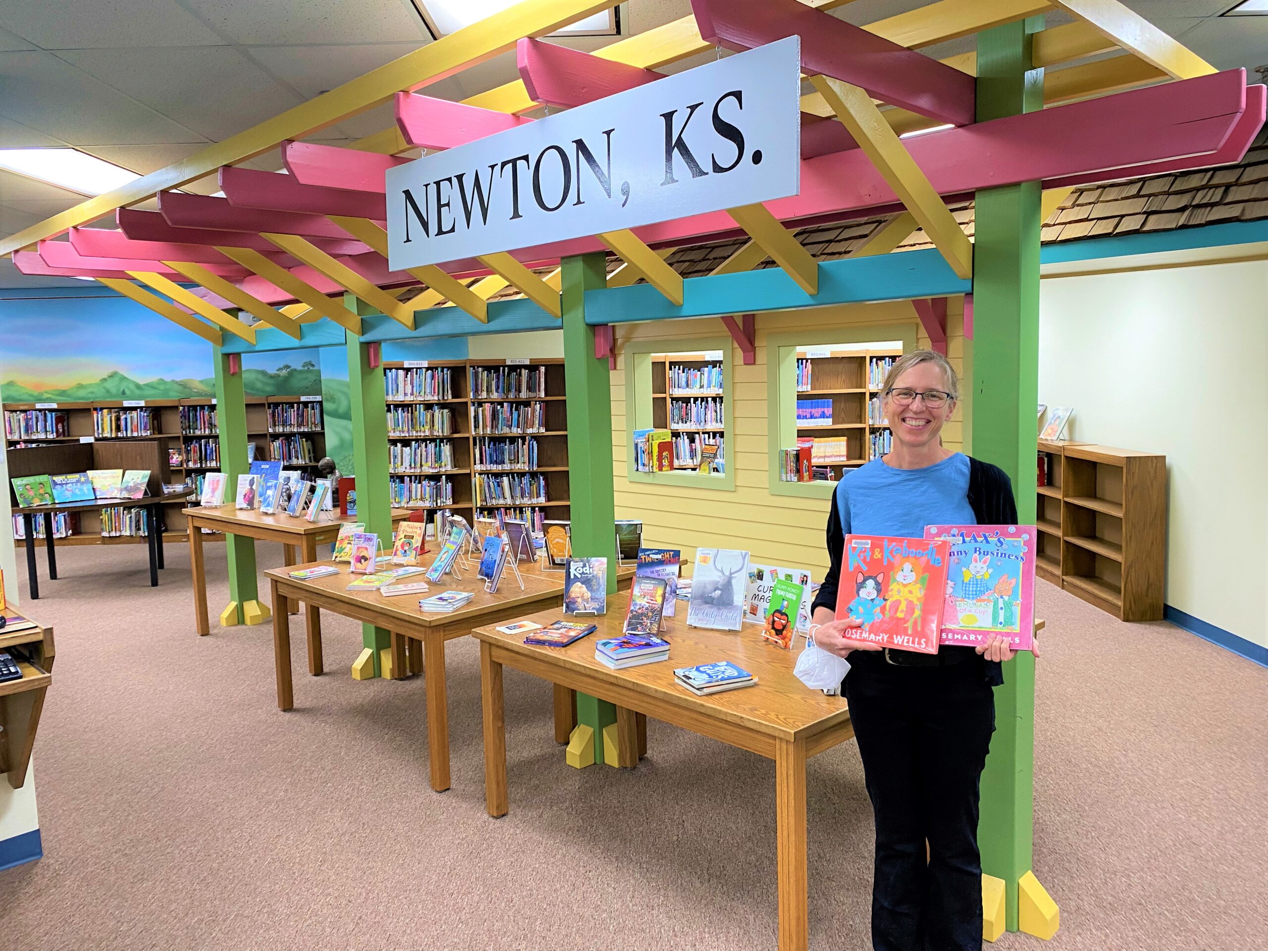 Sara Rickard poses for a photo in the children's department at Newton Public Library. She holds two children's books and stands in front of a background of bookshelves and displays.