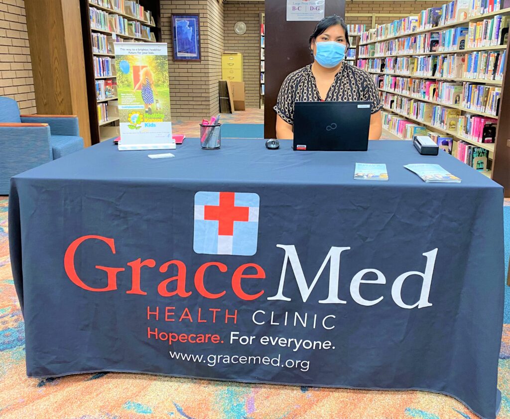 A woman wearing a face mask sits at a table with a GraceMed logo inside Newton Public Library, ready to offer assistance with Medicaid enrollment.