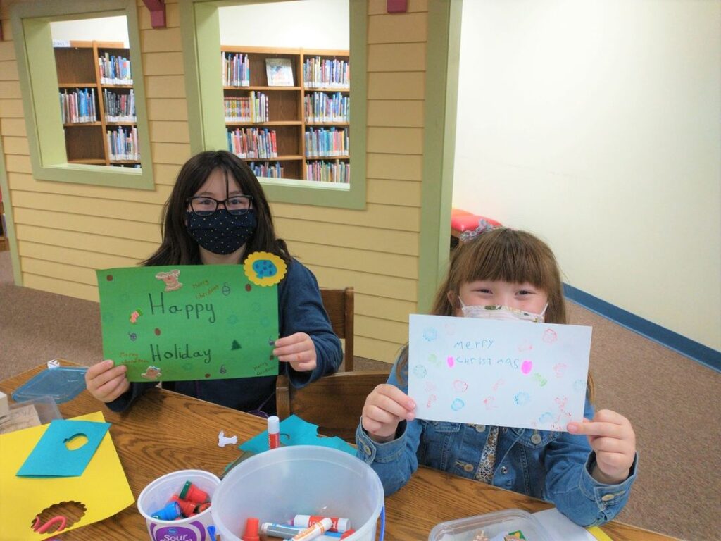 Two young girls wearing masks hold up hand-drawn holiday cards. The children are sitting in the Youth Services level of Newton Public Library.