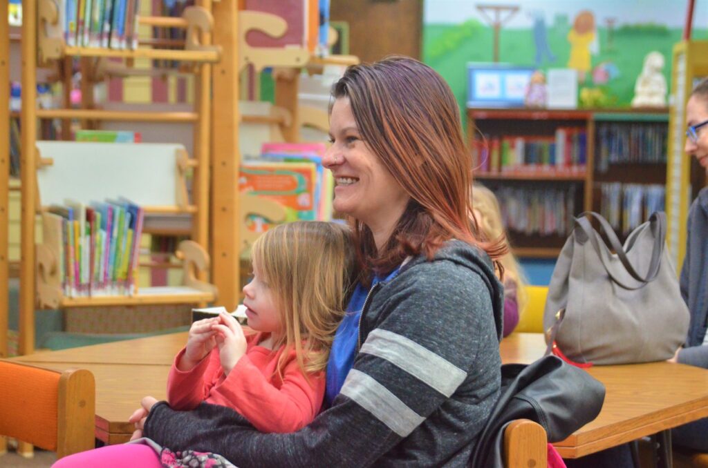 A smiling woman participates in StoryTime with her daughter sitting on her lap.