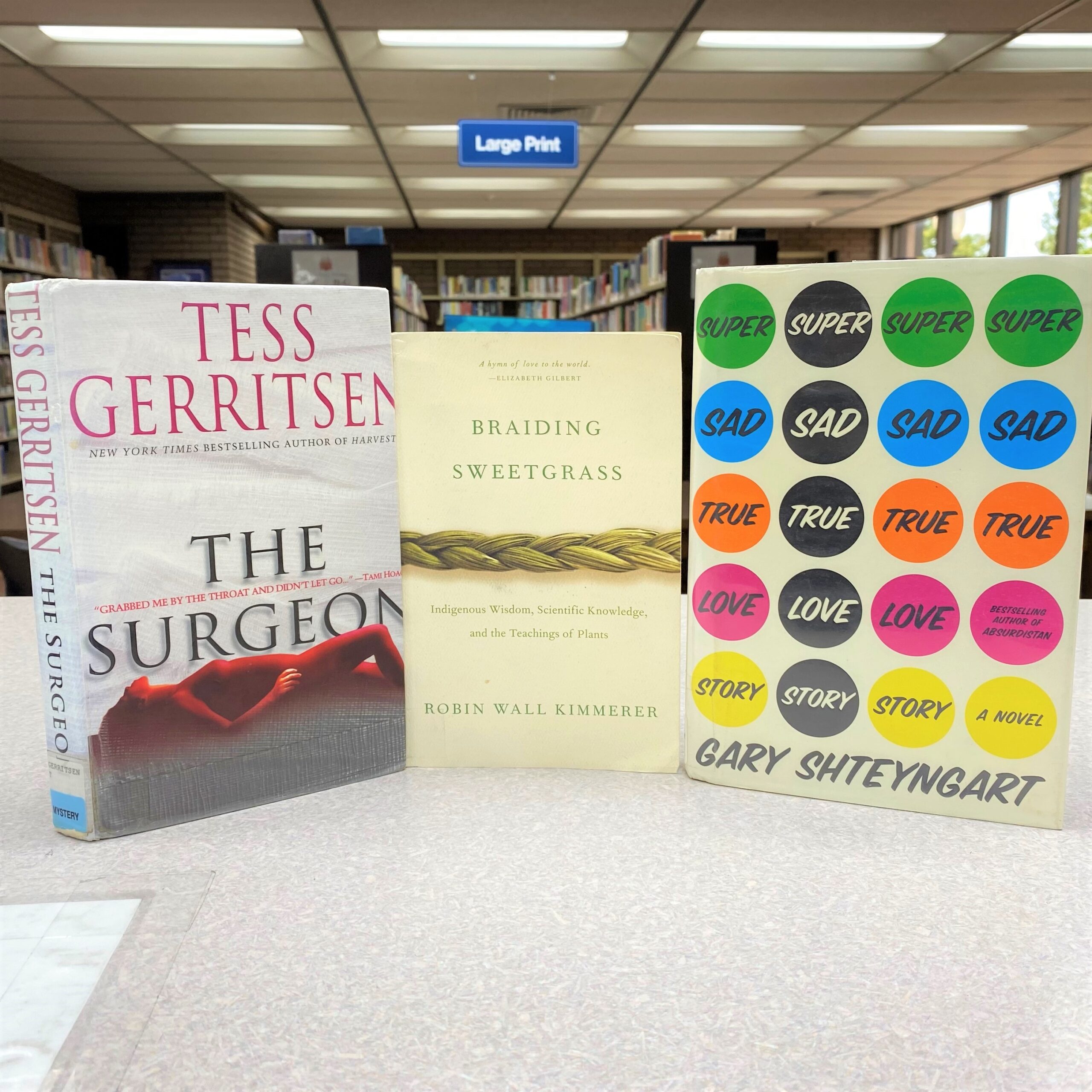 Covers of "The Surgeon," "Braiding Sweetgrass," and "Super Sad True Love Story are pictured sitting on a table.