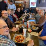 Smiling volunteers enjoy a pizza meal around a long table at a restaurant.