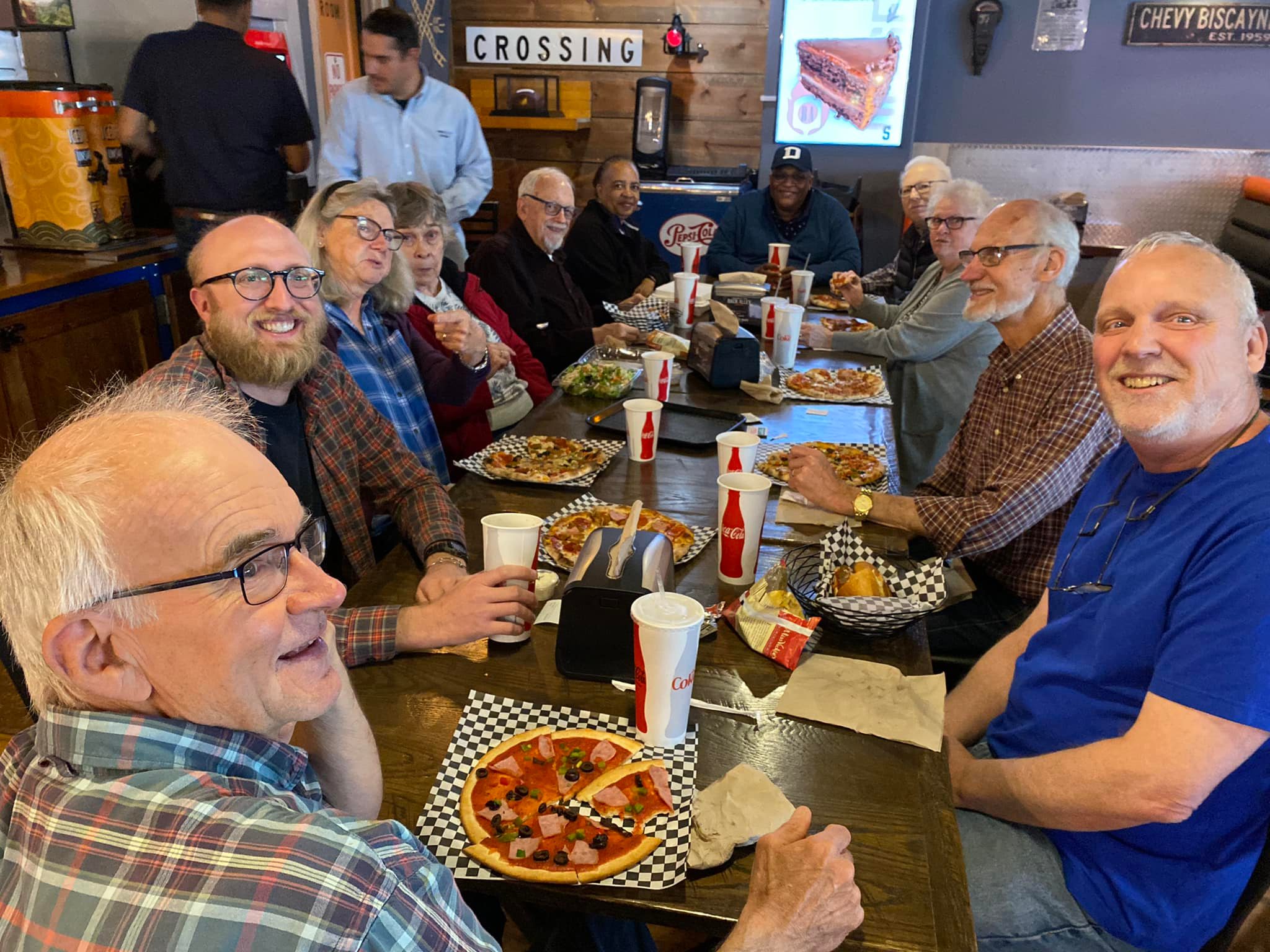 Smiling volunteers enjoy a pizza meal around a long table at a restaurant.