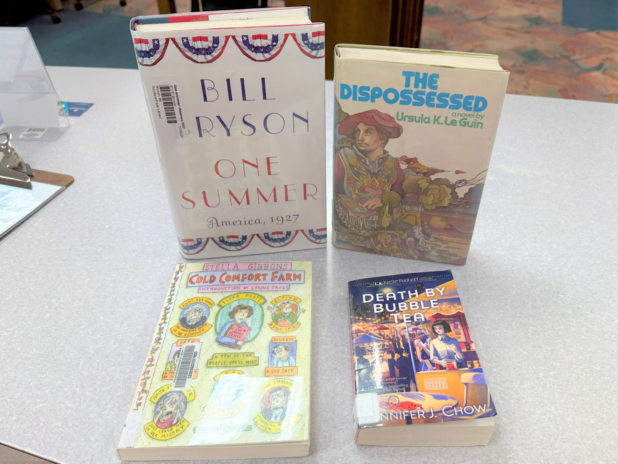 Four books are displayed on a counter: "The Dispossessed," "Cold Comfort Farm," "One Summer: America, 1927," and "Death by Bubble Tea."