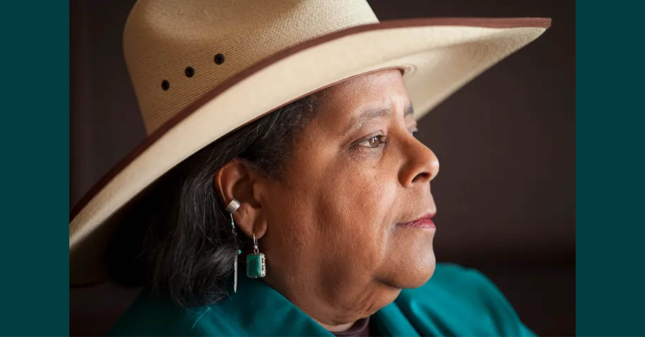 Portrait of Angela Bates. Bates is a Black woman wearing a broad-brimmed hat, a turquoise jacket, and dangling metallic earrings.